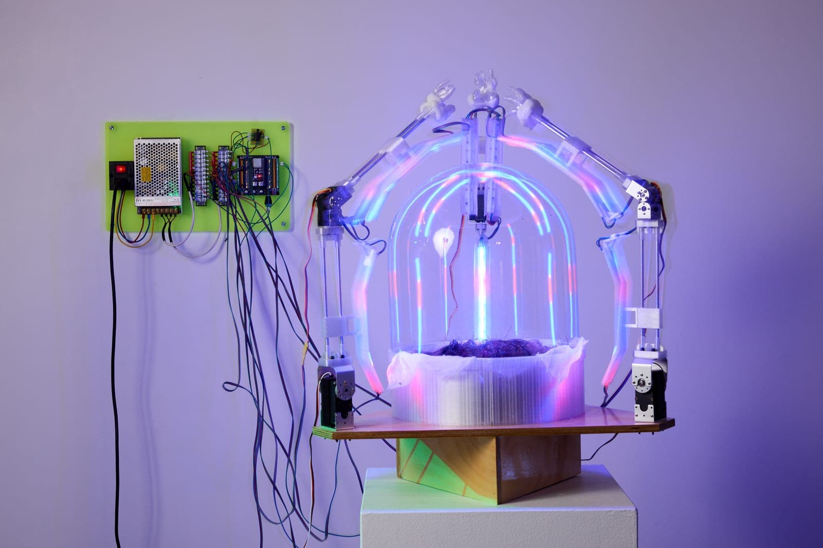 A robotic dome of articulated arms and iridescent light arcs over a nest of wool on a pedestal