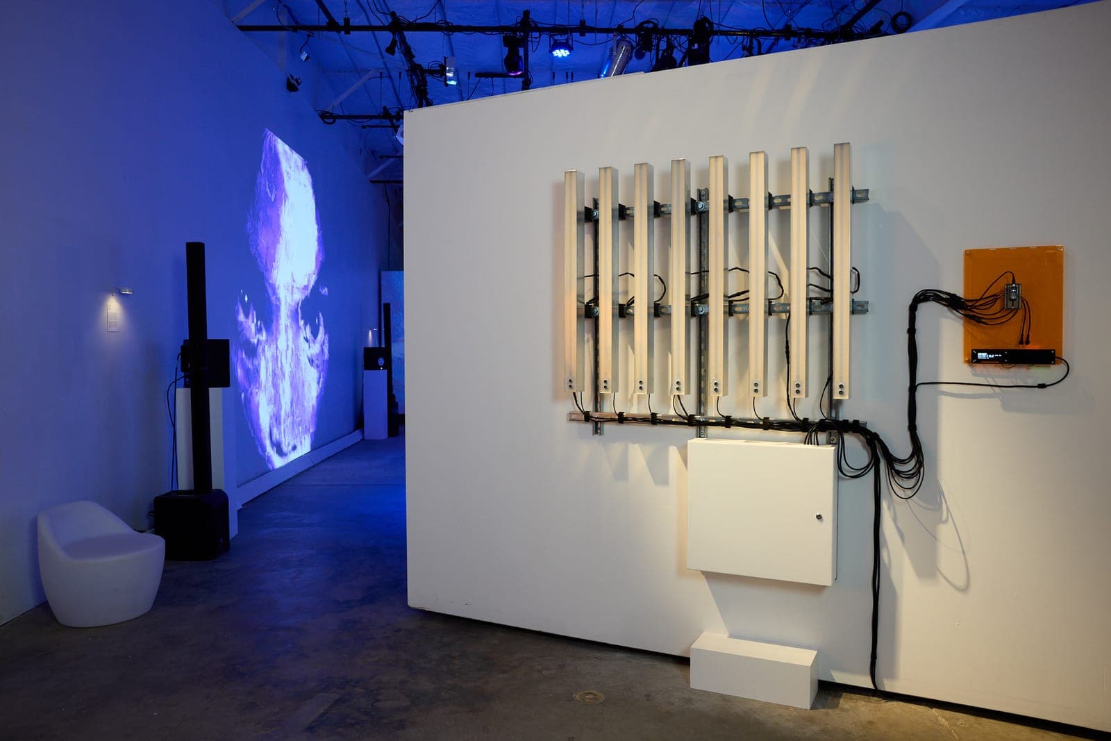 A wall of motorized chime bars wired to an orange controller, a glitched projected face glowing blue in the gallery beyond