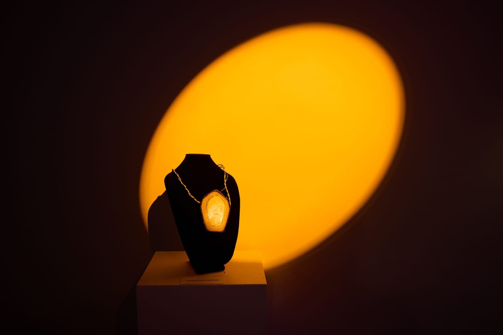A glowing pendant on a black display bust, caught in a warm amber spotlight