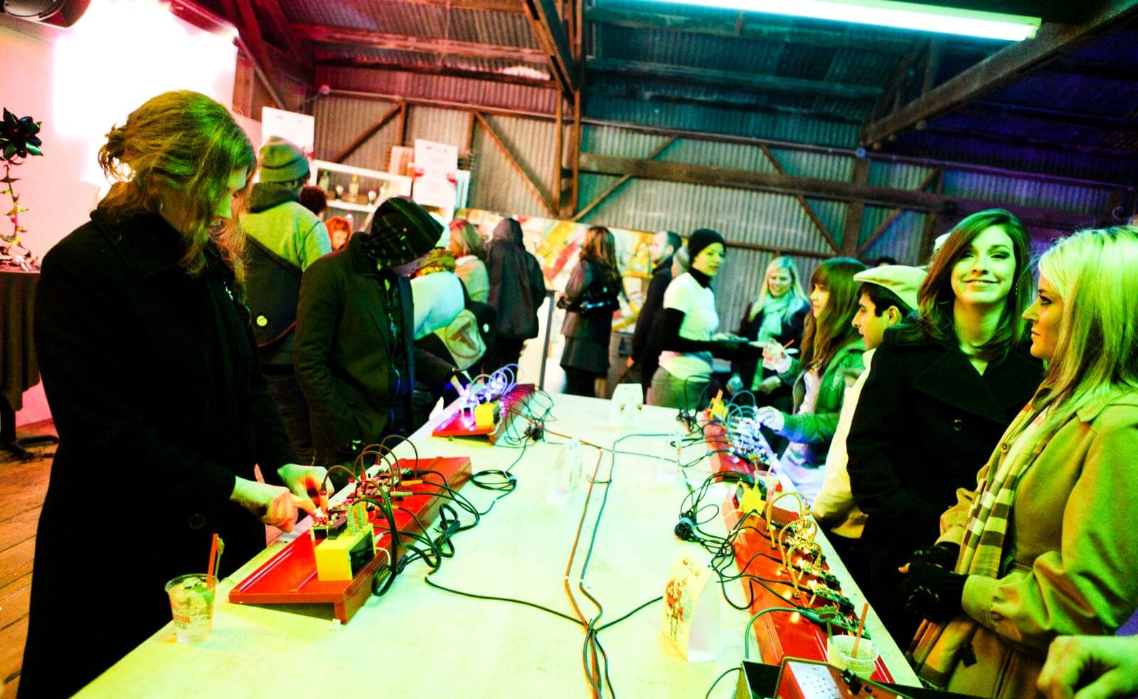 Hands-on circuit workshop in a warehouse space - long table lined with red DIY synth and electronics kits and tangles of patch wire
