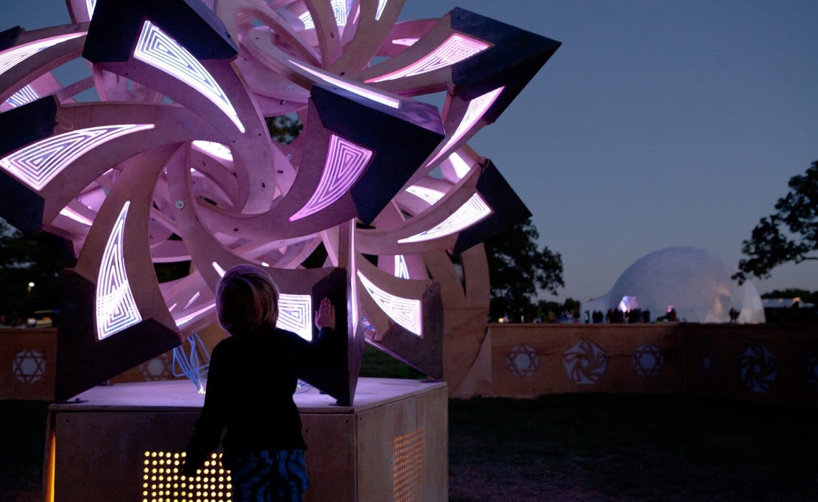 A child reaches up to touch the illuminated frabjous star at dusk, its interlocking plywood curves glowing violet; geodesic dome on the horizon