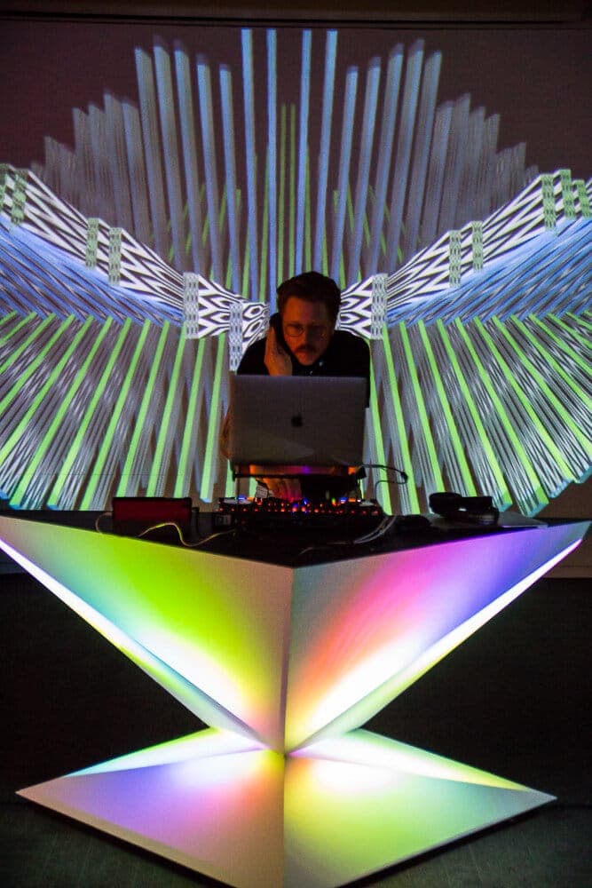 Performer at a glowing geometric podium, a fan of white projected rays spreading across the wall behind