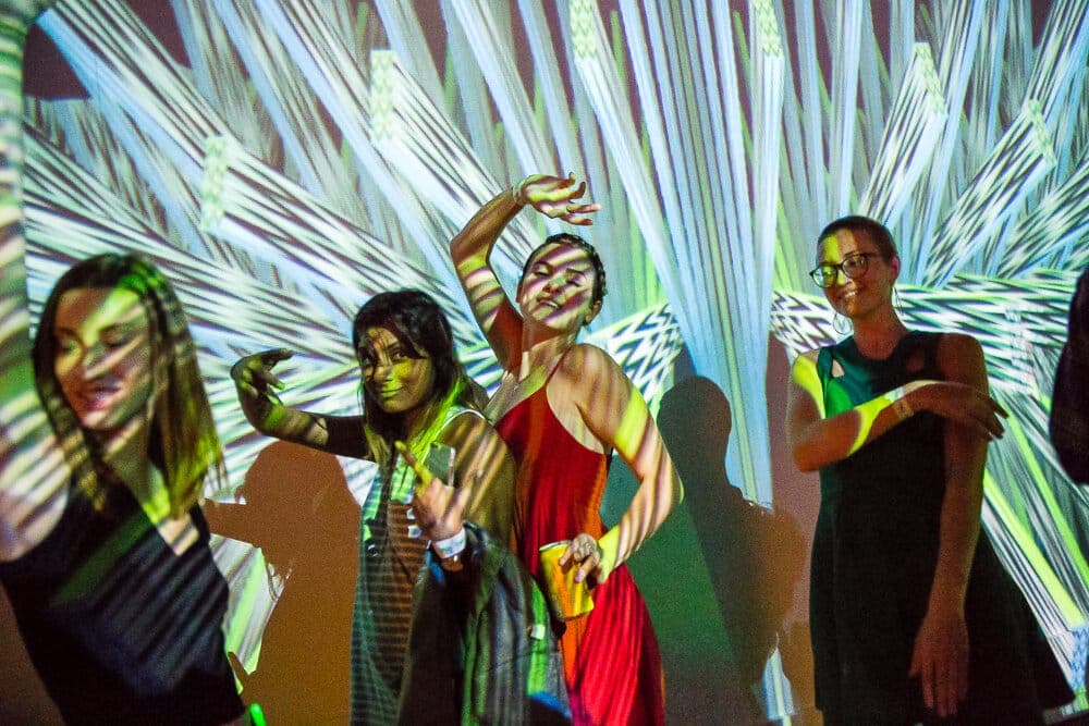 Partygoers dancing with arms raised, striped generative projections raking across them