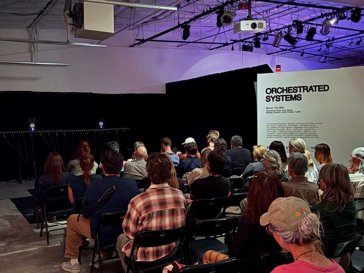 An audience seated before the Orchestrated Systems title wall during an artist talk