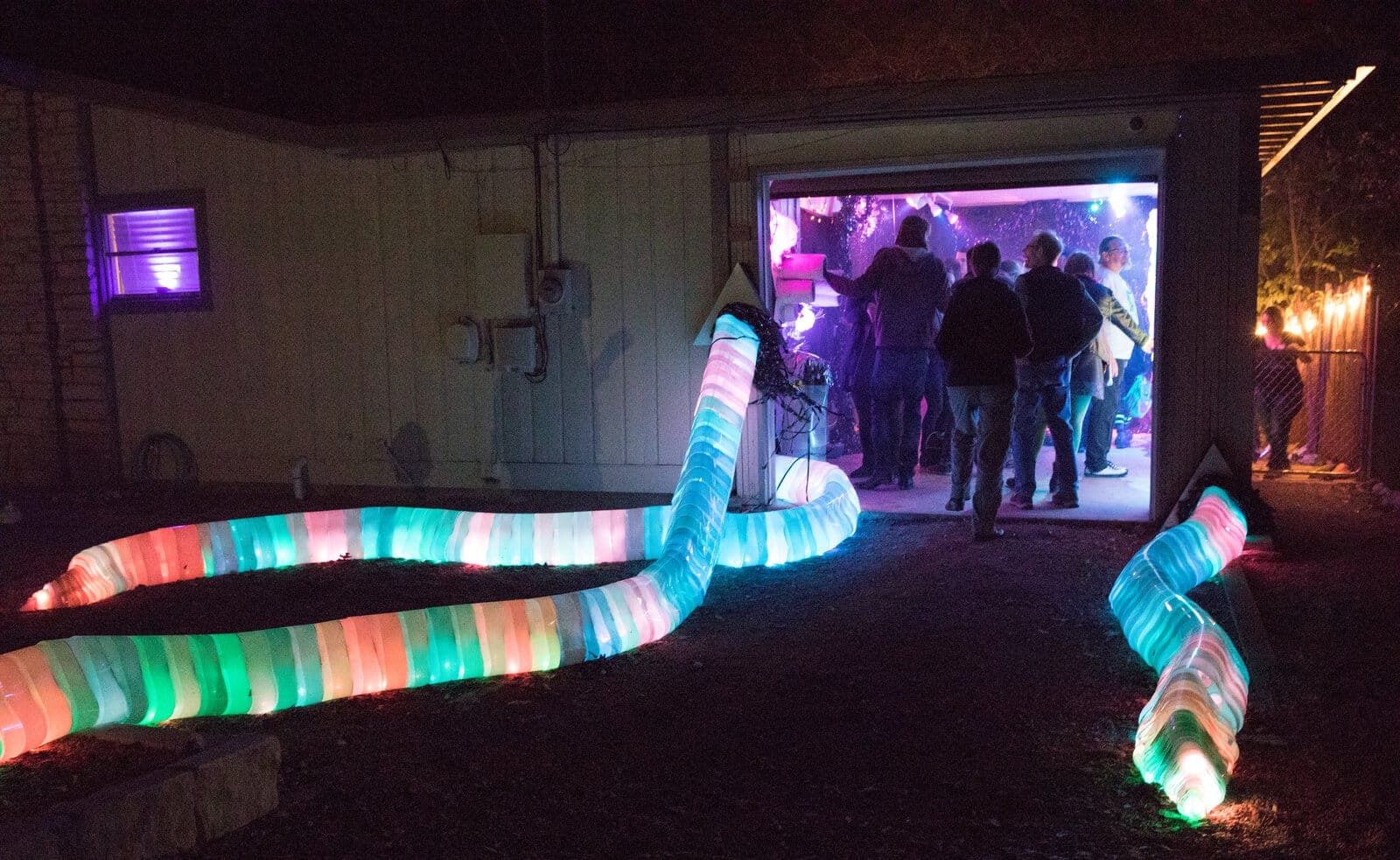 A rainbow-lit segmented tentacle snakes out of the garage doorway into the night as visitors gather inside